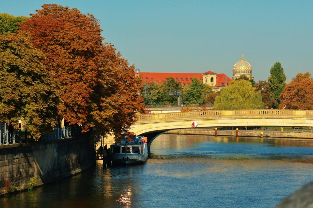 Ein Bild einer Brücke über einen Fluss, mit einem Boot darunter. An der Uferseite stehen Bäume mit herbstlichen Blättern, und im Hintergrund ist ein Gebäude mit rotem Dach und einer goldenen Kuppel zu sehen. Ausblick auf das Social Media Marketing in Deutschland 2025: Solche idyllischen und landschaftlichen Szenen können in Zukunft im deutschen Social - Media - Marketing genutzt werden, um die Attraktivität des Landes zu vermitteln.