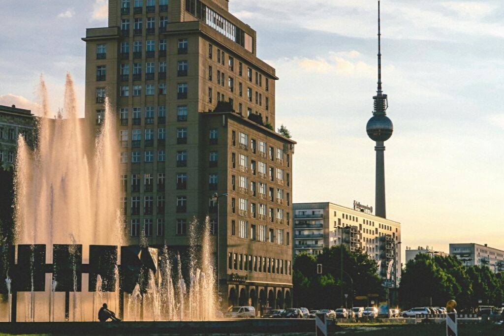 In diesem Bild ist eine beeindruckende Stadtansicht von Berlin zu sehen. Im Vordergrund sprüht ein Wasserspiel hoch, bei dem die Sonnenstrahlen das Wasser in einem warmen Glanz erstrahlen lassen. Ein einzelner Mensch sitzt nearby und genießt die Szene. Dahinter erhebt sich ein imposantes, mehrstöckiges Gebäude mit zahlreichen Fenstern. Im Hintergrund dominiert der Fernsehturm von Berlin, ein Wahrzeichen der Stadt. Xing ist das ideale Netzwerk für deutsche Unternehmen. Mit Bildern wie diesem können Unternehmen die Vibranz und das Potenzial der deutschen Städte zeigen. Sie können auf Xing Stories oder Beiträge erstellen, in denen sie die Verbindung zwischen ihrem Geschäft und der urbanen Kultur Deutschlands herstellen. Indem sie interessante Inhalte wie Fakten über die Stadt oder die Rolle ihres Unternehmens in der Region teilen, können sie die Aufmerksamkeit anderer Fachleute und potenzieller Geschäftspartner auf Xing gewinnen und ihr Netzwerk erweitern.